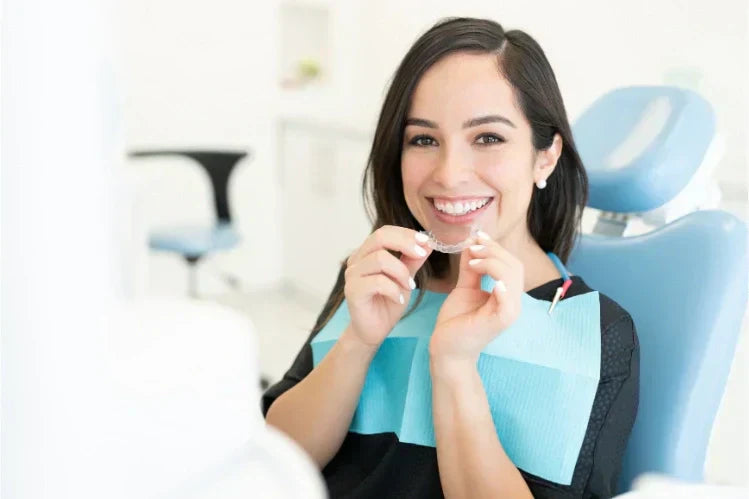 Una mujer en el consultorio de un dentista, sosteniendo un alineador y sonriendo.